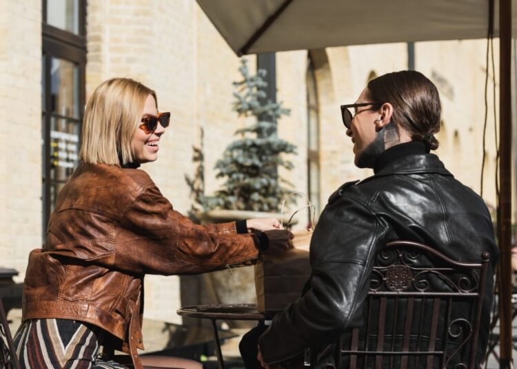 cheerful blonde woman in sunglasses holding paper bag while sitting on metallic bistro chair near
