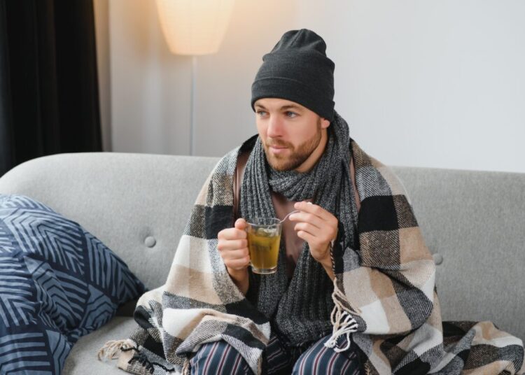 It's cold at home in wintertime Man freezing in his house in winter because of broken thermostat young guy wrapped in woolen plaid shivering while sitting on sofa in living room interior