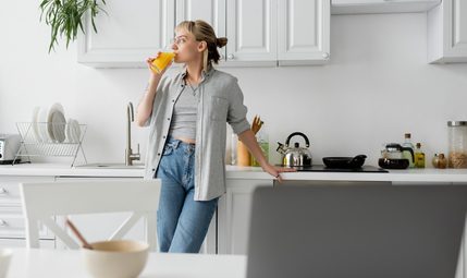 tattooed woman in eyeglasses drinking orange juice and standing near kitchen worktop next to desk with devices bowl with cornflakes and cup of coffee with saucer at home freelance lifestyle